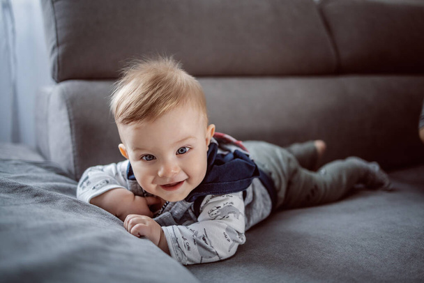 Wesoły szczęśliwy mały chłopiec uśmiecha się i leży na brzuchu na kanapie w salonie. - Zdjęcie, obraz