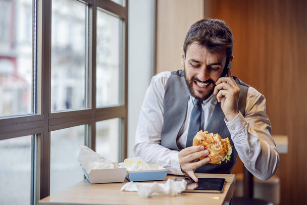 Young handsome bearded businessman sitting in fast food restaurant, having phone call and eating burger. - Photo, Image