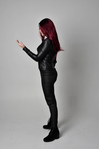 Volledig portret van meisje met rood haar in zwart leren jasje, broek en laarzen. Stand in zijprofiel, geïsoleerd tegen een grijze studioachtergrond. - Foto, afbeelding