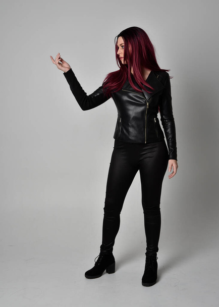 Full length portrait of  girl with red hair wearing black leather jacket, pants and boots. Standing pose  in side profile, isolated against a grey studio background. - Photo, image