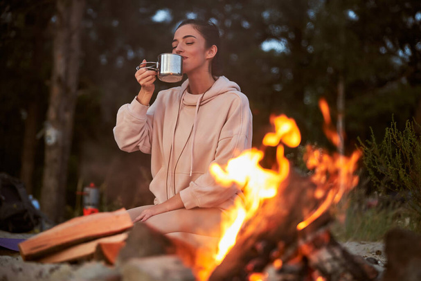 Beautiful young woman drinking tea near campfire - Foto, Imagen