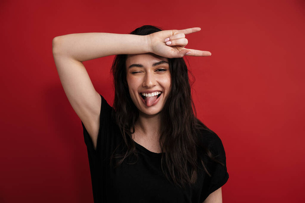 Photo of brunette funny woman in casual t-shirt winking and showing horn sign isolated over red background - Photo, Image