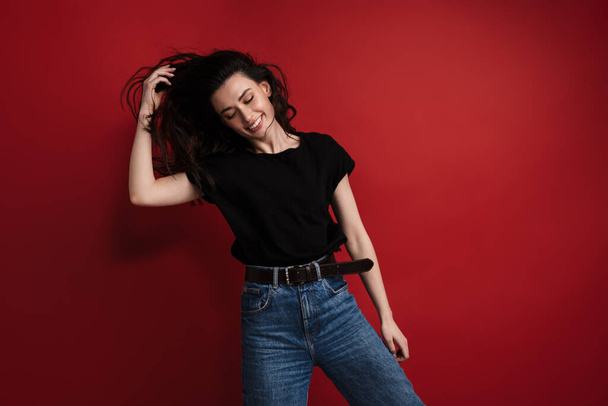 Photo of brunette cheerful woman in casual t-shirt laughing and dancing isolated over red background - Foto, Imagen