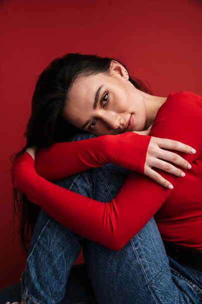 Photo of brunette beautiful woman sitting on chair and looking at camera isolated over red background - Фото, зображення