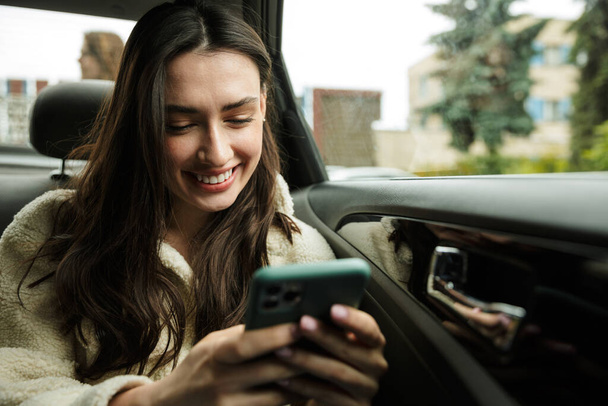 Beautiful smiling young brunette woman wearing coat sitting on a back of a car, using mobile phone - Fotografie, Obrázek