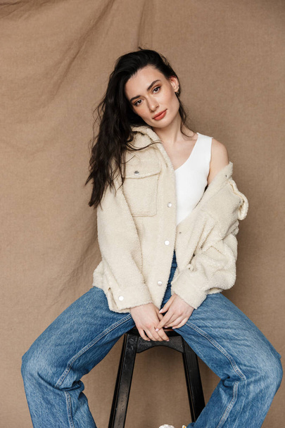 Photo of beautiful young woman in jacket looking at camera while sitting on chair isolated over beige background - Fotó, kép