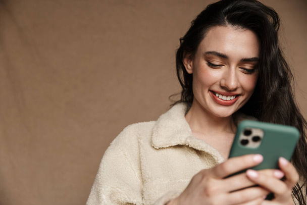 Photo of beautiful young woman in jacket smiling and holding cellphone isolated over beige background - Zdjęcie, obraz