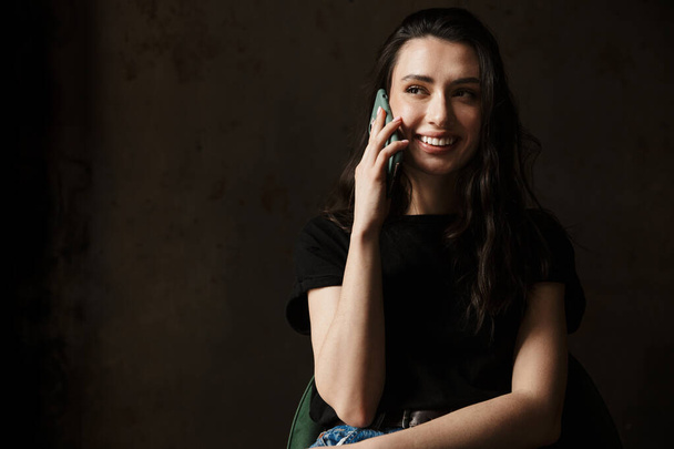 Photo of smiling young woman sitting on chair and talking on cellphone isolated over dark background - Zdjęcie, obraz