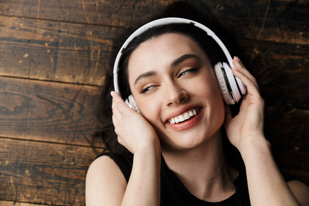 Photo from above of beautiful young brunette woman wearing headphones smiling and lying on wooden floor - Foto, Imagem