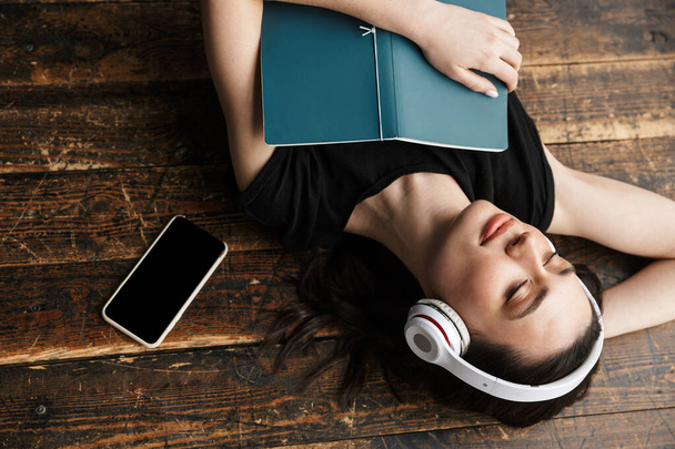 Photo from above of beautiful young brunette woman wearing headphones lying on wooden floor with book and cellphone - Фото, зображення