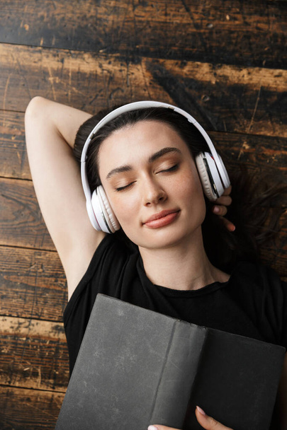 Photo from above of beautiful young brunette woman wearing headphones lying on wooden floor with book - Foto, immagini