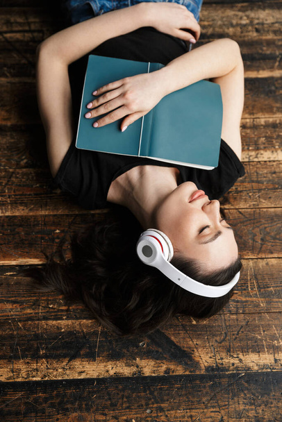 Fotografie z výše krásné mladé brunetky žena na sluchátka ležící na dřevěné podlaze s knihou - Fotografie, Obrázek