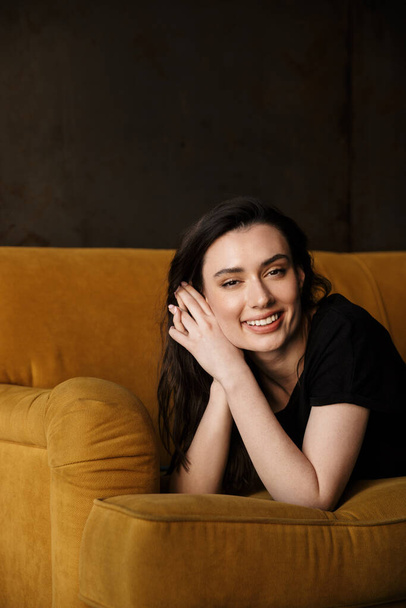 Photo of beautiful young brunette woman smiling and lying on yellow couch in modern apartment - 写真・画像