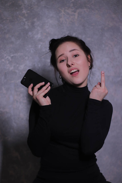 Emotionales Porträt eines brünetten Mädchens in schwarzem Rollkragen mit schwarzem Telefon, zeigt Mittelfinger gegen graue Wand - Foto, Bild
