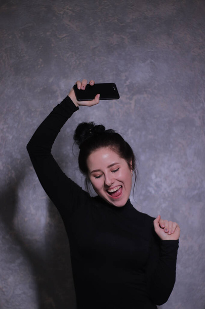 érzelmi portré egy boldog barna lány egy fekete garbó tartó fekete telefon egy szürke fal - Fotó, kép
