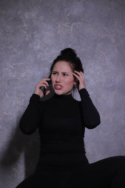 érzelmi portré egy barna lány egy fekete garbó méltatlankodva beszél telefonon egy szürke fal - Fotó, kép