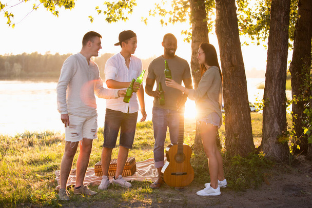 Skupina přátel cinkajících během pikniku na pláži pivními skleničkami. Životní styl, přátelství, zábava, víkend a odpočinek koncept. - Fotografie, Obrázek