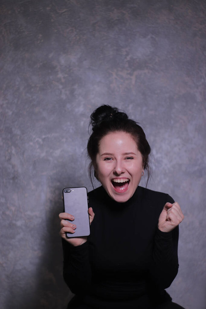 érzelmi portré egy barna lány egy fekete garbó tartó fekete telefon egy szürke fal - Fotó, kép