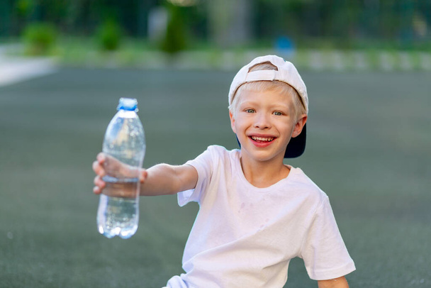 портрет блондина в спортивной форме, сидящего на зеленой лужайке на футбольном поле и держащего бутылку воды. Обучение детей - Фото, изображение