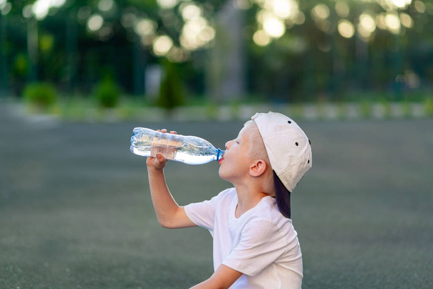 Porträt eines blonden Jungen in Sportuniform, der auf einem grünen Rasen auf einem Fußballplatz sitzt und Wasser aus einer Flasche trinkt, Sportteil. Ausbildung der Kinder, Freizeit der Kinder. - Foto, Bild