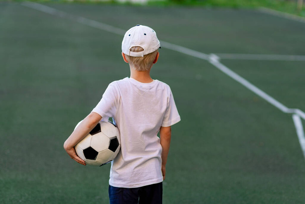 Futbol sahasında yeşil çimlerde oturan spor elbiseli bir çocuk futbol topu, arka görüş, spor sayfası, antrenman. - Fotoğraf, Görsel
