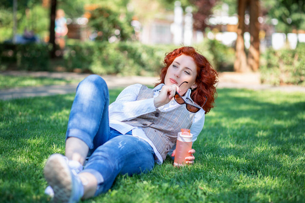 Sevimli kızıl saçlı bir kız parkta çimlerin üzerinde yatıyor ve bir şeyler düşünüyor ya da hayal ediyor. Bir yaz parkında elinde bir şişe suyla yürüyen bir kadın. - Fotoğraf, Görsel