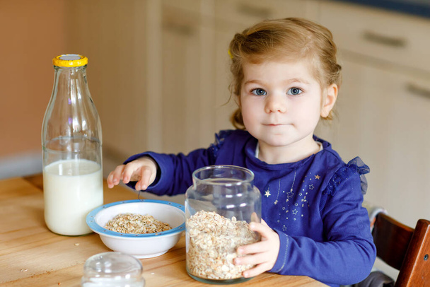 愛らしい幼児の女の子は朝食のためにミルクと健康的なオートミールを食べています。キッチンに座って、オート、シリアルを準備するのが楽しいカラフルな服のかわいい幸せな赤ちゃん。家の中で - 写真・画像