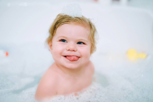 Милая очаровательная малышка, принимающая пенную ванну в ванной. Тоддлер играет с резиновыми игрушками. Красивый ребенок веселится с красочными игрушками жевательной резинки и пеной пузырьков - Фото, изображение