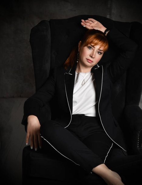 A wealthy, bossy woman in a black trouser suit sits in a high chair in a comfortable position and looks at you. Cropped shot with dark gray background. - Photo, image