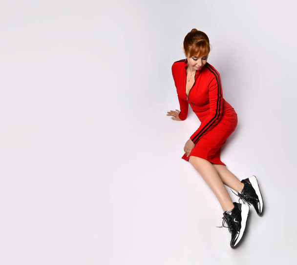 Top view of beautiful woman in red slim-line dress sitting on floor in relaxed pose looking at her black sneakers. Full length shot isolated on white - Φωτογραφία, εικόνα