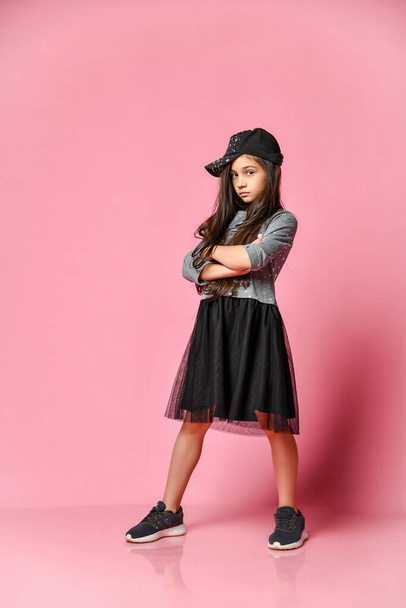 Portrait of a little hipster girl in a dark dress with a baseball cap and sneakers, on a pink background - Φωτογραφία, εικόνα