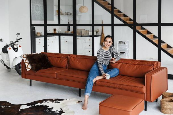 Attractive smiling young asian woman wearing casual clothes relaxing on a leather couch at home, having cup of coffee, using mobile phone - Фото, изображение