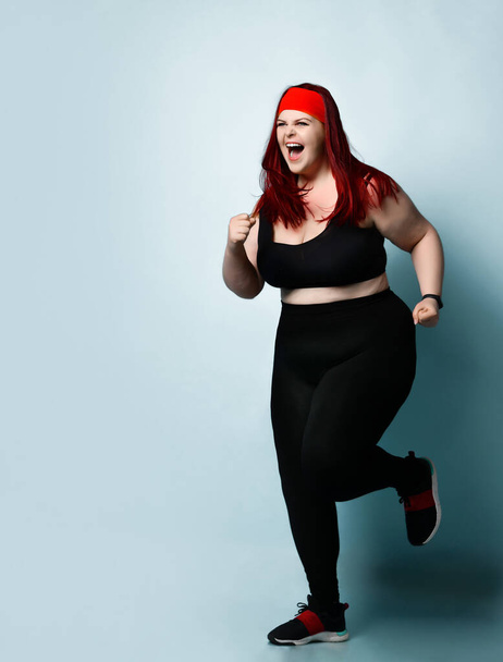 Young plus-size girl with red hair in black sportswear shouts with anger and expression keeping fists clenched on blue background. Copy space - Foto, afbeelding