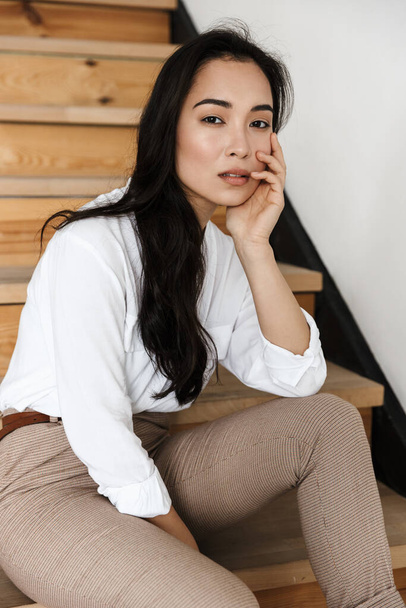 Sonriente atractiva joven asiática mujer usando camisa blanca sentado en una escalera en casa - Foto, imagen