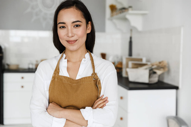 Smiling attractive young asian woman wearing apron standing at the kitchen at home - Photo, Image