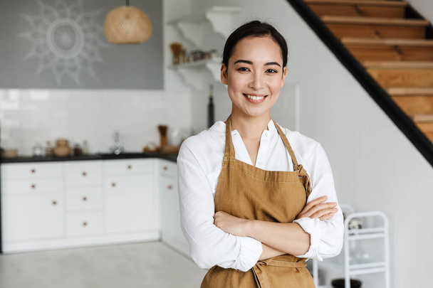 Smiling attractive young asian woman wearing apron standing at the kitchen at home - Fotografie, Obrázek