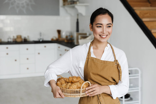 Smiling attractive young asian woman wearing apron standing at the kitchen at home, holding basket with fresh croissants - Fotoğraf, Görsel