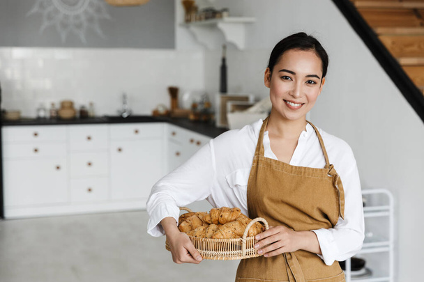 Hymyilevä houkutteleva nuori aasialainen nainen yllään esiliina seisoo keittiössä kotona, pitäen koria tuoreilla croissanteilla - Valokuva, kuva