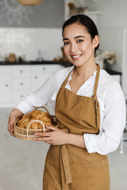 Smiling attractive young asian woman wearing apron standing at the kitchen at home, holding basket with fresh croissants - Foto, afbeelding