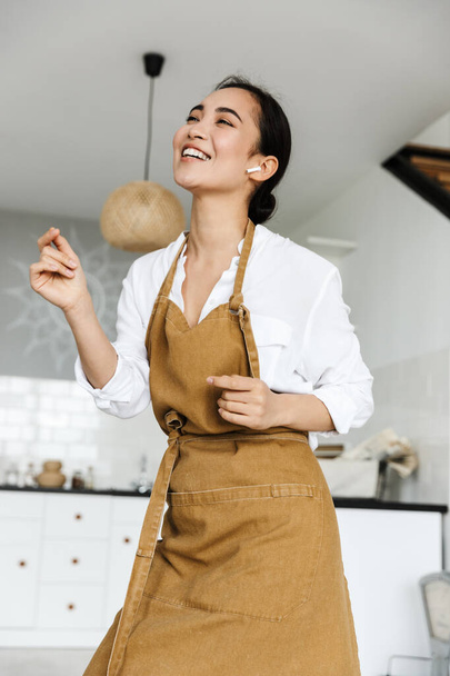 Cheerful young asian woman wearing apron dancing while listening to music with wireless earphones in the kitchen - Foto, afbeelding