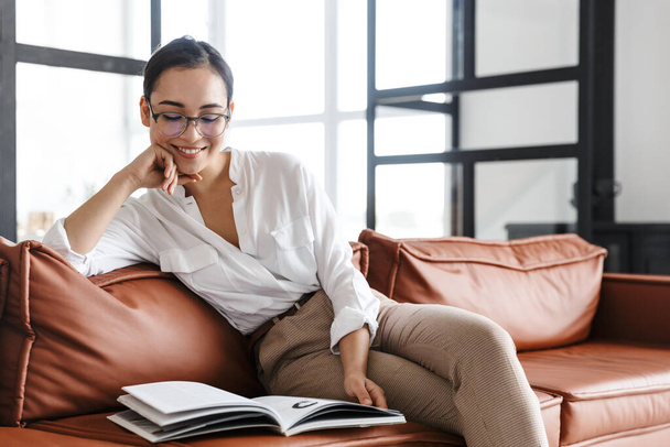 Attractive smiling young asian business woman relaxing on a leather couch at home, reading a magazine - Photo, image