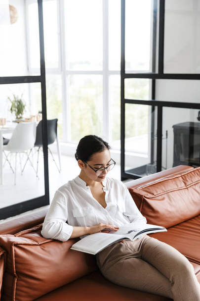 Attractive smiling young asian business woman relaxing on a leather couch at home, reading a magazine - Fotó, kép