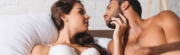 Vue panoramique de la femme en soutien-gorge toucher le menton du petit ami musclé sur le lit  - Photo, image