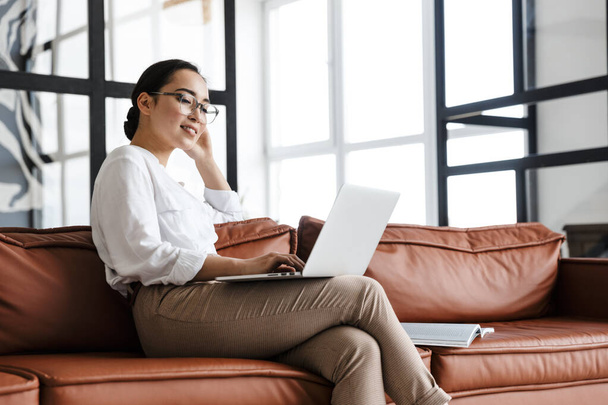 Приваблива усміхнена молода азіатська бізнес-леді розслабляється на шкіряному дивані вдома, працює на ноутбуці
 - Фото, зображення