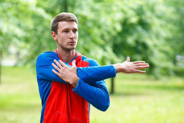 Front view of a sportsman stretching hands while standing outdoor in a park and looking by side. Close up cropped photo. - Foto, afbeelding