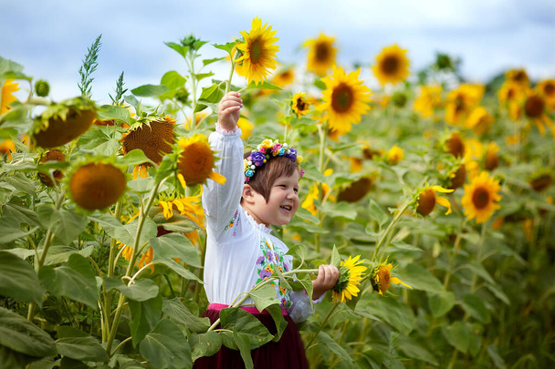 Feliz niña ucraniana sonriente con una camisa bordada y una corona de flores con una bandera amarilla y azul en sus manos en un campo de girasoles. Día de la Independencia de Ucrania. Día de la bandera nacional - Foto, imagen