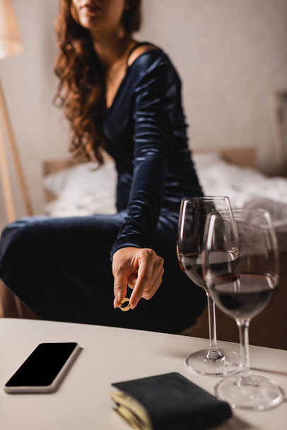 Ausgeschnittene Ansicht einer jungen Frau, die Ehering auf Couchtisch in der Nähe von Weingläsern, Smartphone und Geldbörse im Schlafzimmer platziert  - Foto, Bild