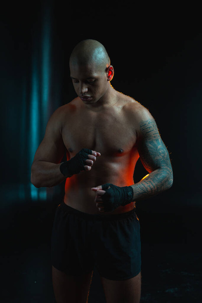 Serious muscular tattooed man in black shorts standing alone in the darkness and wrapping boxing bandages around the wrist - Foto, Bild