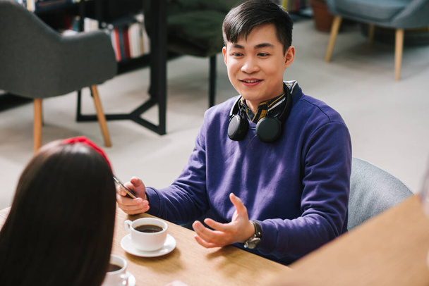 Cheerful man sitting with a smartphone in hand and a cup of coffee on the table in front of him and smiling while talking - 写真・画像