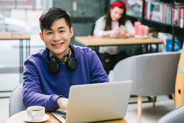 Positive Asian young man sitting at the table with a modern laptop and a cup of coffee and smiling. Young lady on the background - Фото, зображення